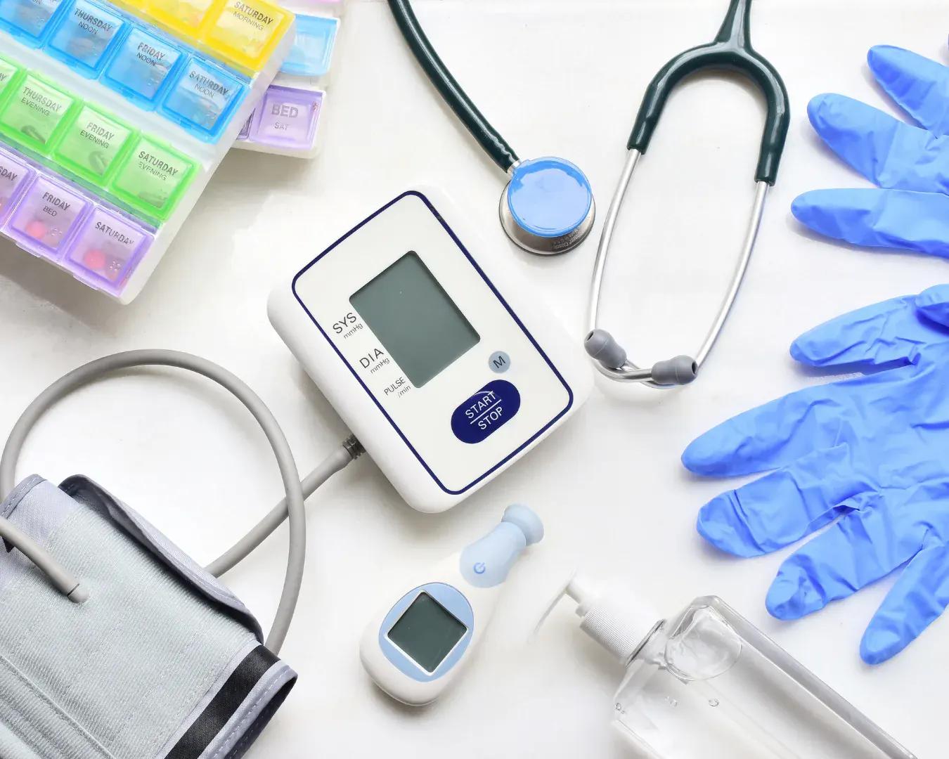 Medical supplies on a white surface: blood pressure monitor, stethoscope, pill organizer, gloves, thermometer, and hand sanitizer