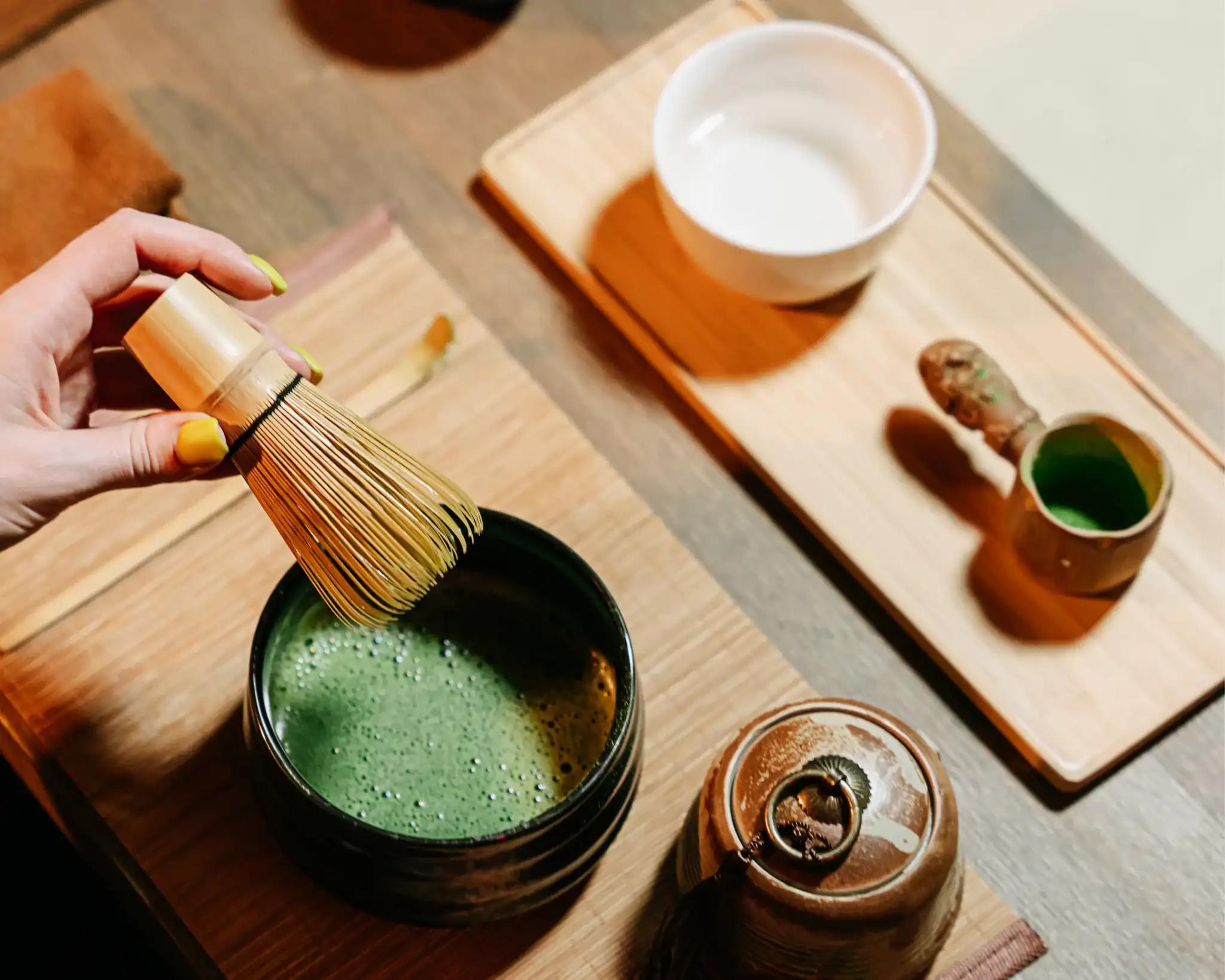 Hands whisking frothy matcha in a ceramic bowl with bamboo tools—artisanal tea preparation