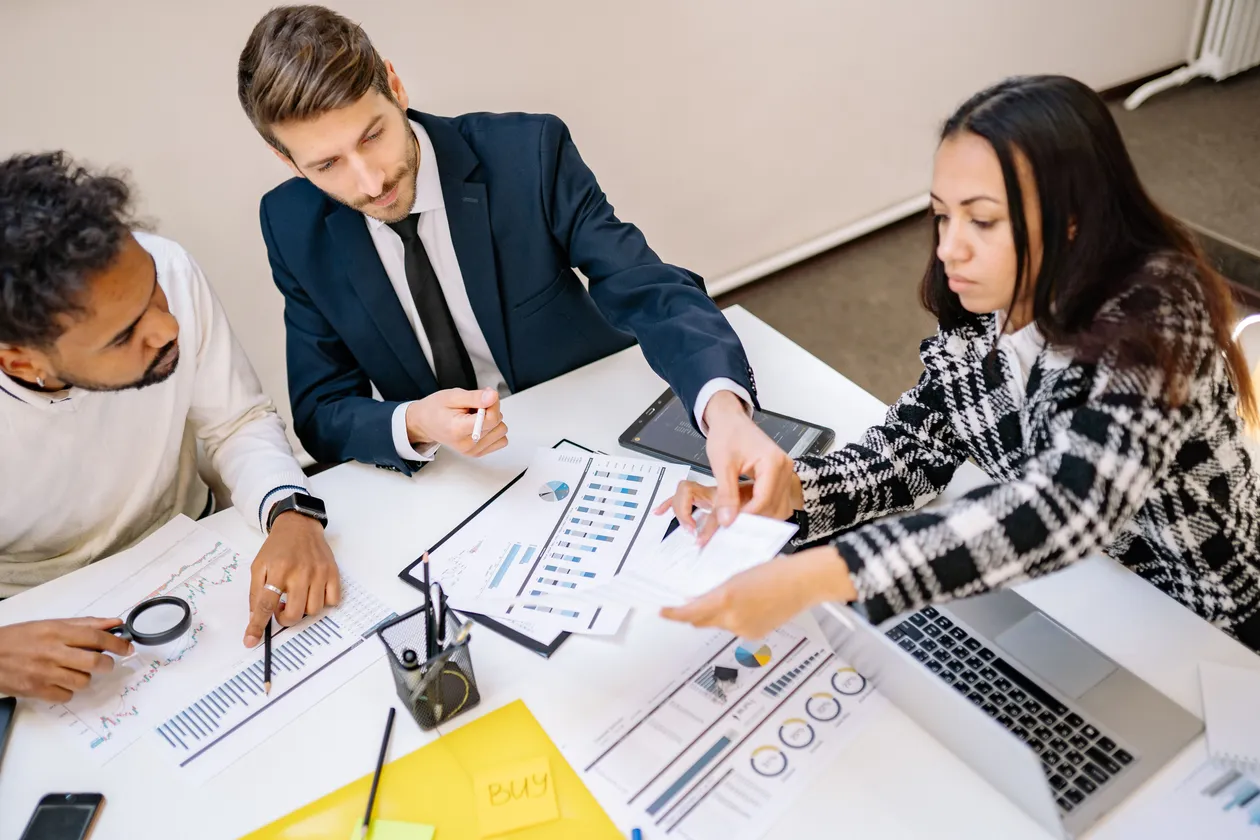 Three colleagues in a meeting reviewing printed bar charts, spreadsheets, and a laptop laid out across a white table