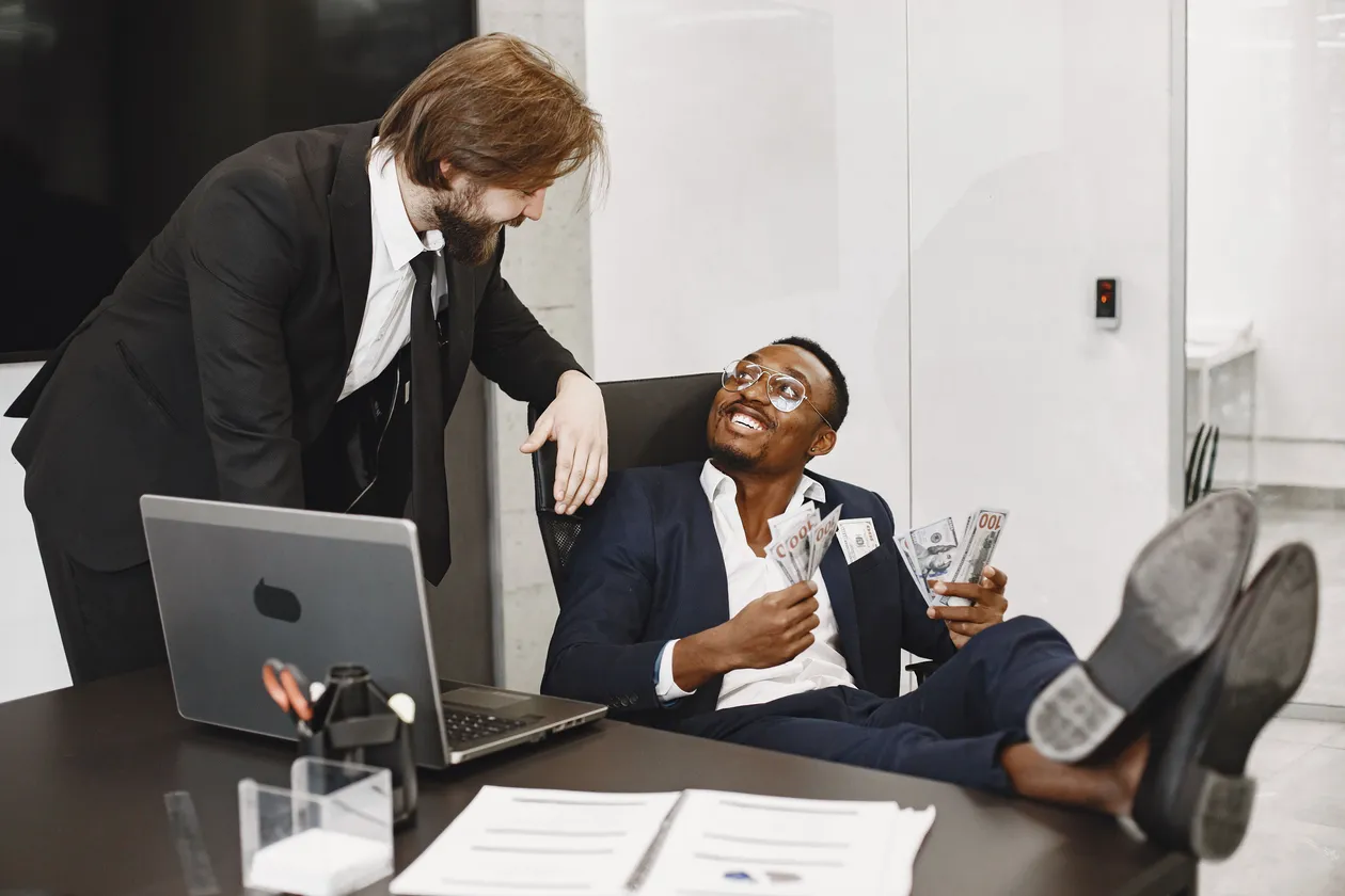 Two colleagues in suits at a desk with a laptop, one seated leaning back playfully holding fanned-out banknotes while the other stands beside him