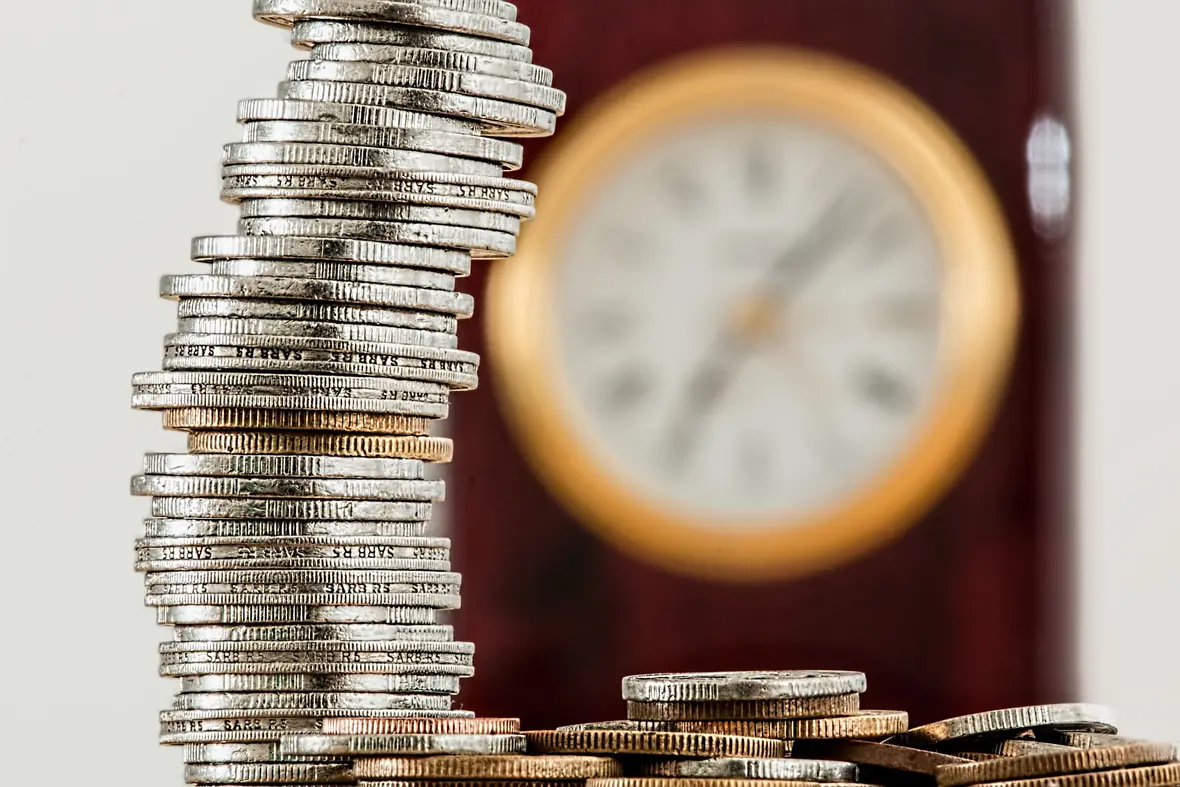 Tall stack of silver coins next to a blurred antique wall clock