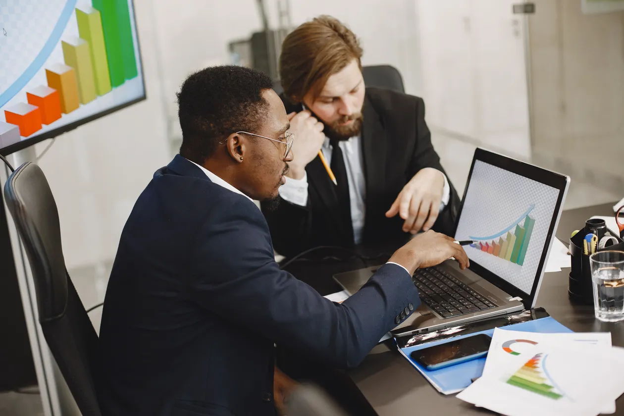 Two men in suits reviewing a rising bar chart on a laptop at an office desk with colorful graphs behind them
