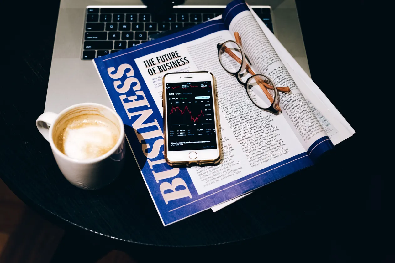 Smartphone showing stock chart on top of an open business magazine beside eyeglasses, a coffee cup, and a laptop