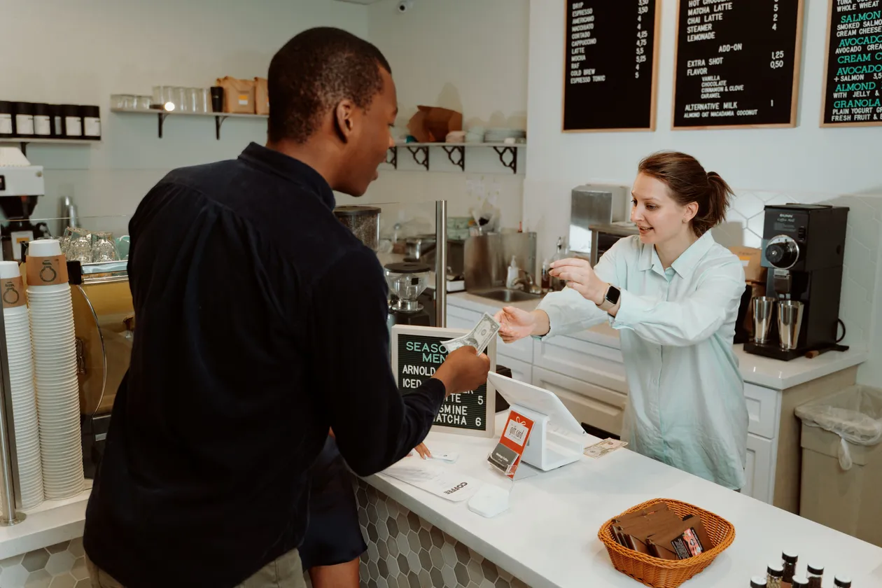 Barista at a cafe counter handing change to a customer beside a small point-of-sale display and menu boards