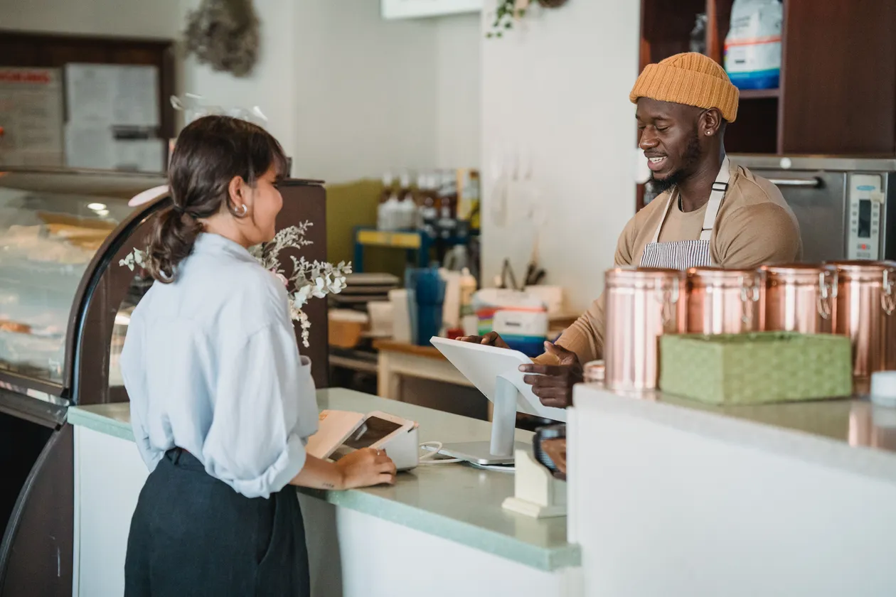 Woman at a cafe counter placing an order with a smiling barista wearing an orange beanie behind copper canisters