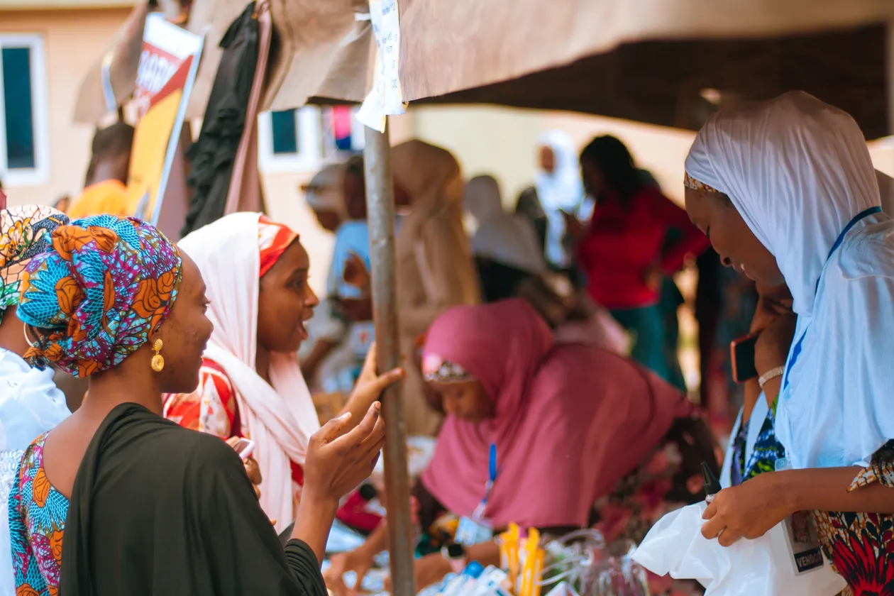 Group of women in brightly colored traditional attire gathered around an outdoor market stall, talking and shopping