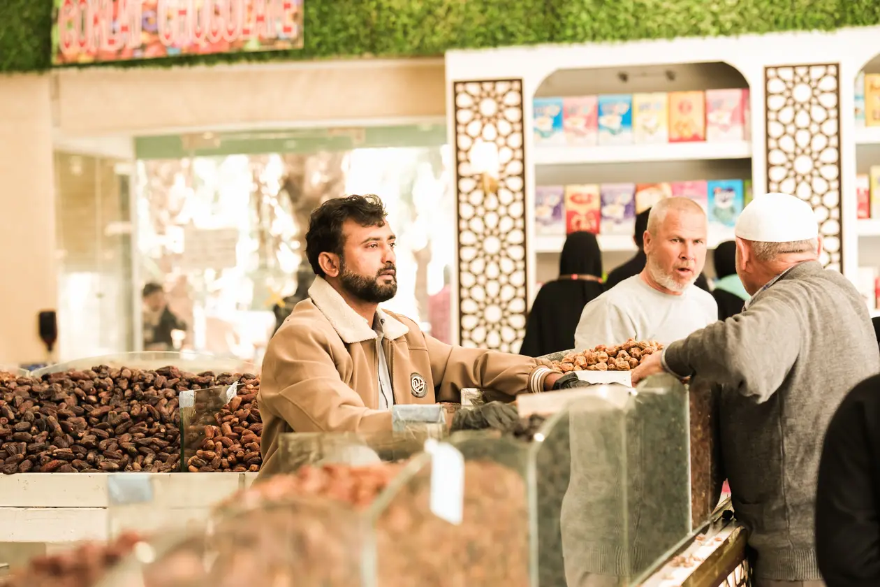 Shoppers browsing a brightly lit date market stall in Saudi Arabia, with the vendor behind trays of dried dates and seasoning packets