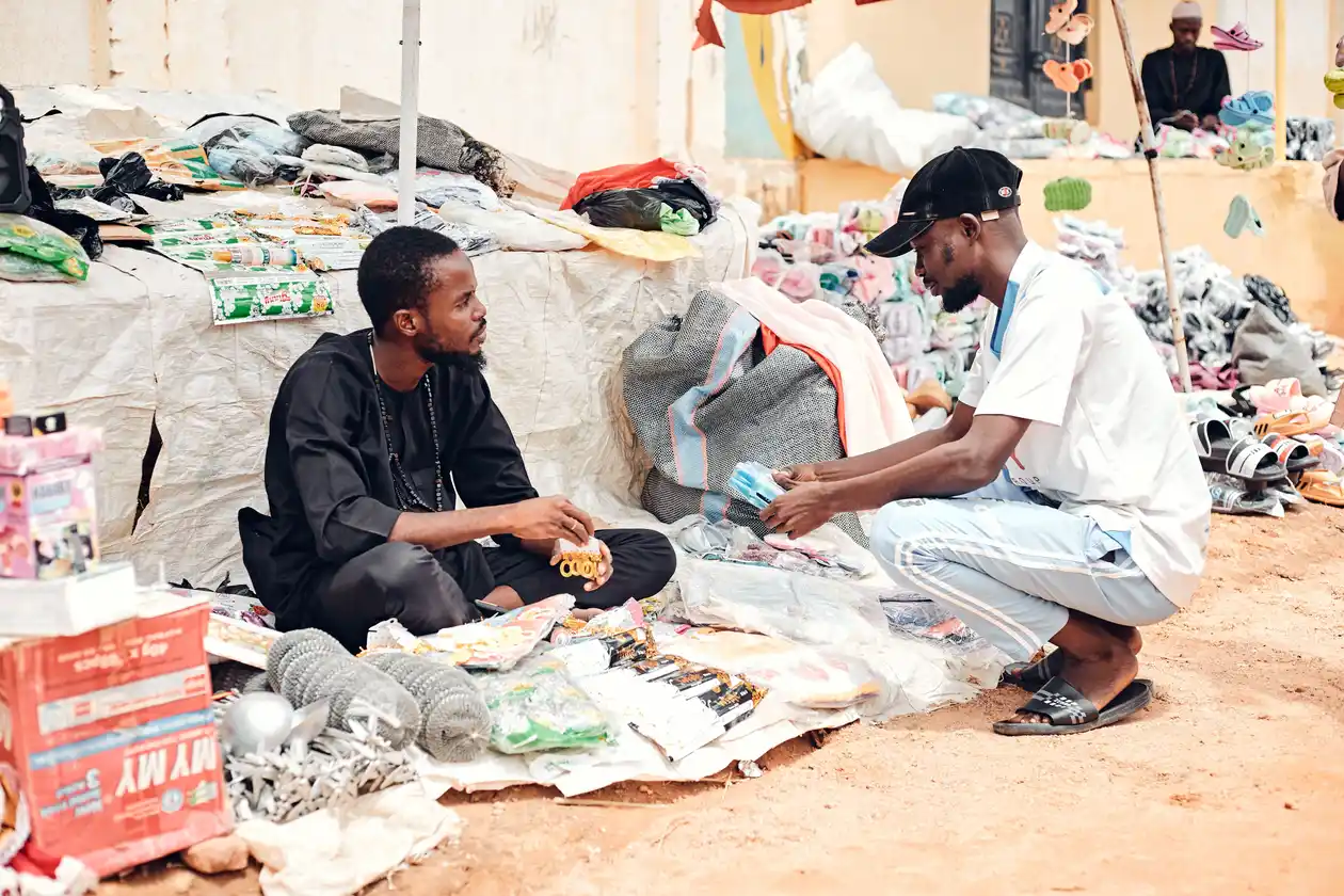 Two vendors sitting on the ground at a street market, surrounded by piles of clothing, household goods, and bags of goods for sale