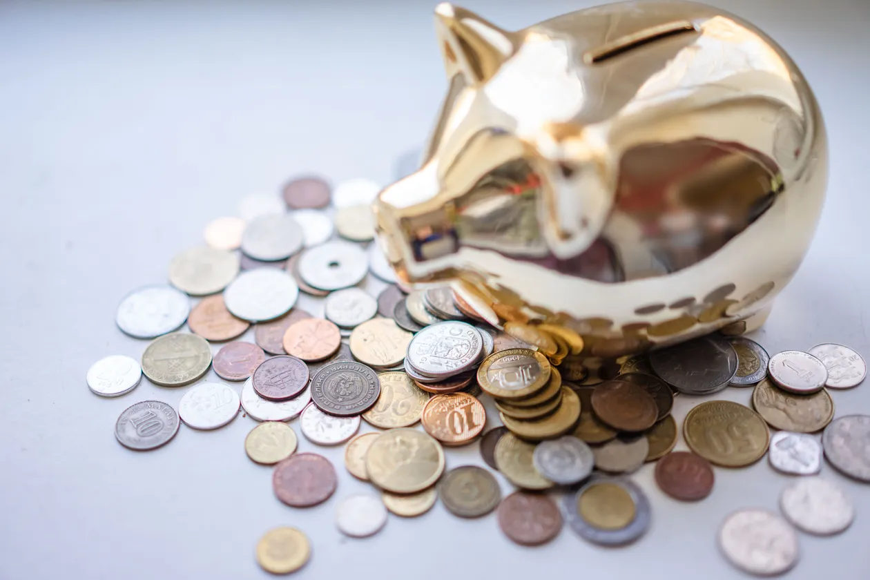 Shiny gold piggy bank surrounded by a pile of scattered silver and gold coins on a light backdrop
