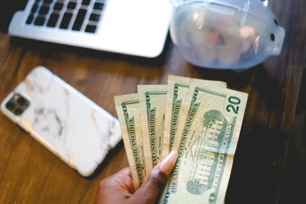 Hand fanning out several U.S. twenty-dollar bills beside a smartphone and a piggy bank on a wooden desk