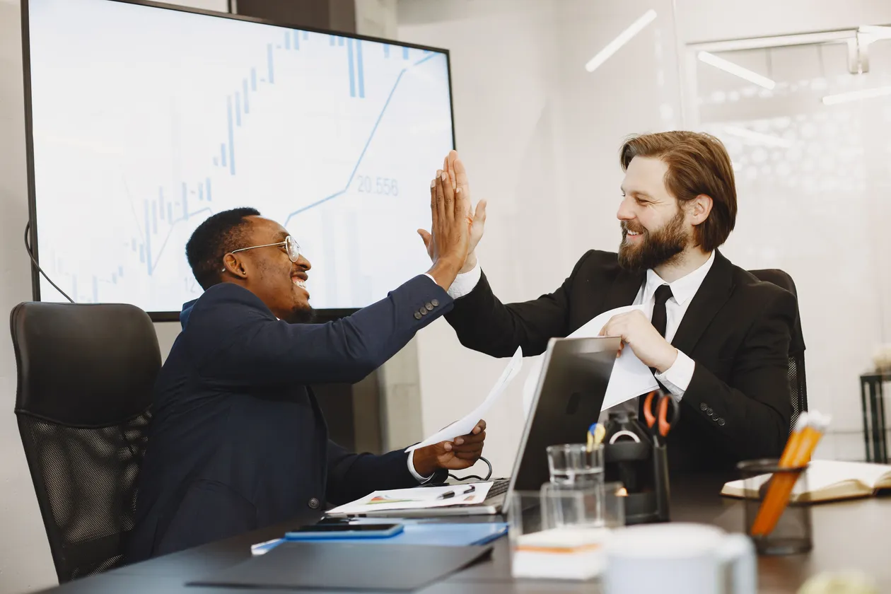Two men in suits giving each other a celebratory high five across a desk with a laptop in a modern meeting room, a chart visible on the wall behind them
