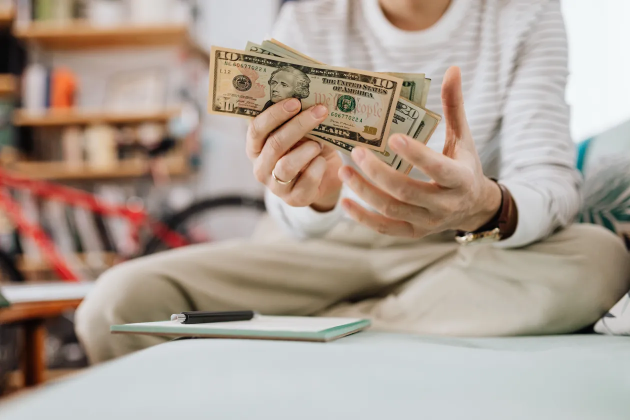 A person sitting cross-legged on a sofa counting a fan of US ten and twenty dollar bills, with a notebook and pen resting beside them