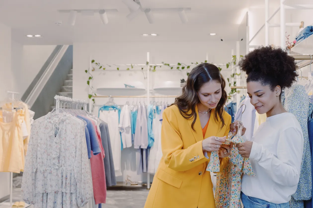 Two women browsing a floral dress together inside a brightly lit retail clothing store with racks of garments on both sides